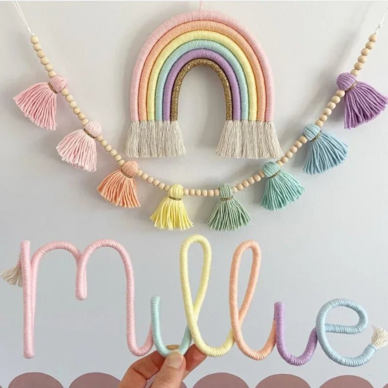 Décoration murale pour chambre d'enfant présentant un arc-en-ciel en macramé aux couleurs pastel et or pailleté, suspendu au-dessus d'une guirlande de perles de bois et de pompons en fil. Une main tient une décoration en fil formant le prénom "Millie", reprenant les mêmes teintes douces. L'ensemble crée une ambiance douce et artisanale.