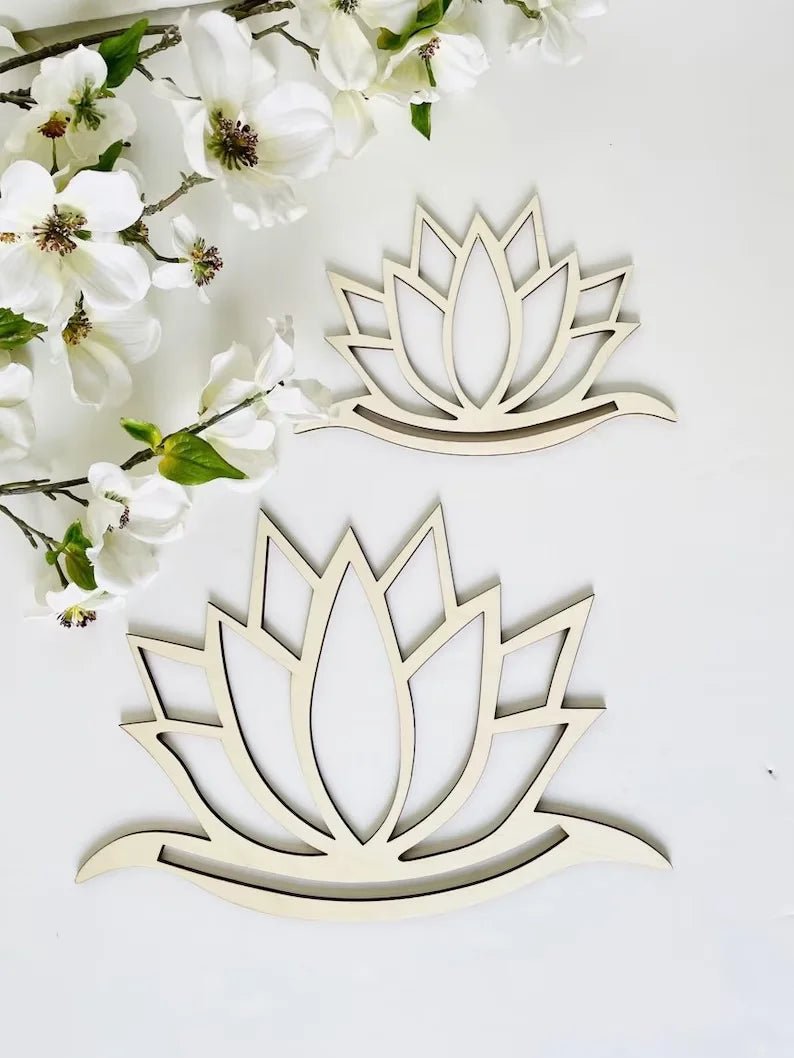 Deux décorations murales en forme de fleur de lotus en bois clair sont posées sur un fond blanc, accompagnées d'une branche fleurie aux délicates fleurs blanches. Ces éléments pourraient être intégrés dans une création en macramé, ajoutant une touche naturelle et spirituelle à l'ouvrage. La simplicité des formes et des couleurs évoque la sérénité.