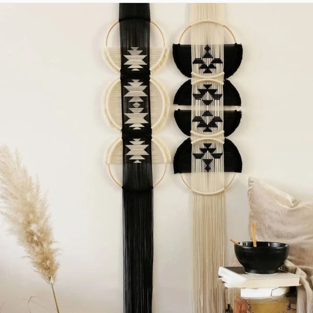 Deux décorations murales en macramé artisanal ornent un mur blanc. L'une, à gauche, est principalement noire avec des motifs géométriques beiges, tandis que l'autre, à droite, inverse les couleurs. Les deux créations suspendues sont composées de cercles en bois et de longues franges de fils.