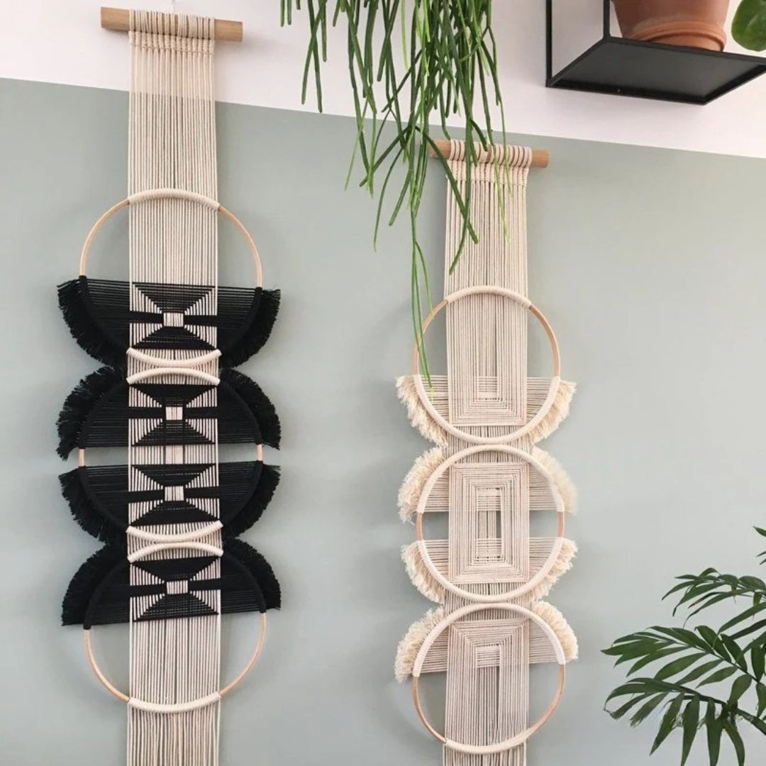 Deux décorations murales en macramé suspendues côte à côte sur un mur gris clair. Celle de gauche est principalement noire avec des motifs blancs, tandis que celle de droite est dans des tons écrus et beiges. Ces créations artisanales apportent une touche bohème et texturée à l'espace.