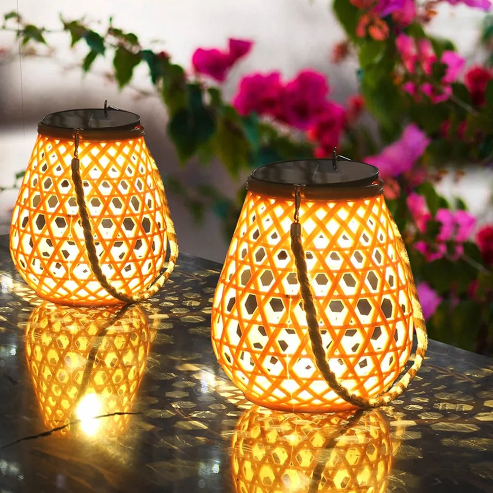 Deux lanternes Lampe solaire jardin haut de gamme-Bambou Tissé émettent une lumière jaune chaude sur une surface brillante, avec des bougainvilliers roses flous en arrière-plan, créant une ambiance extérieure chaleureuse et vibrante.