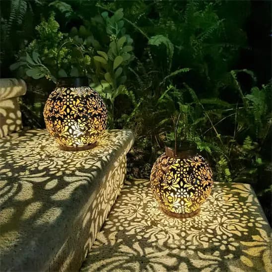Deux lanterne solaire extérieure taupe de Mafrenesiedecoration projettent des ombres de feuilles complexes sur des marches en pierre entourées dune végétation luxuriante. Alimentées par des panneaux solaires, elles diffusent une lueur chaleureuse qui crée une atmosphère chaleureuse et met en valeur le cadre du jardin avec un éclairage extérieur efficace.