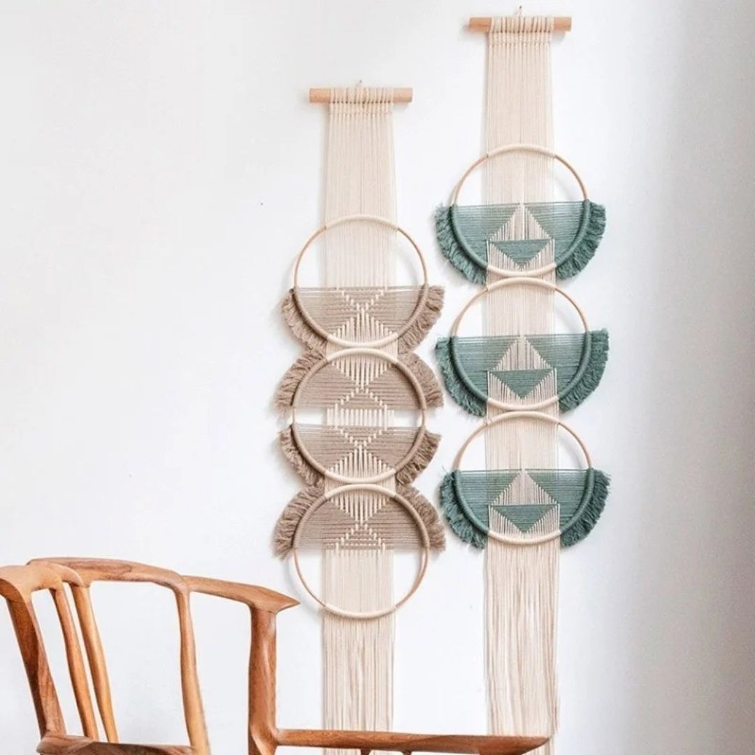 Deux macrame murales ornent un mur blanc, chacune suspendue à un bâton en bois. L'une présente trois cercles décoratifs dans des tons de beige et de crème, tandis que l'autre aligne trois cercles aux motifs géométriques en nuances de vert et de beige. Un fauteuil en bois clair est partiellement visible au premier plan, à gauche. Marcrame.