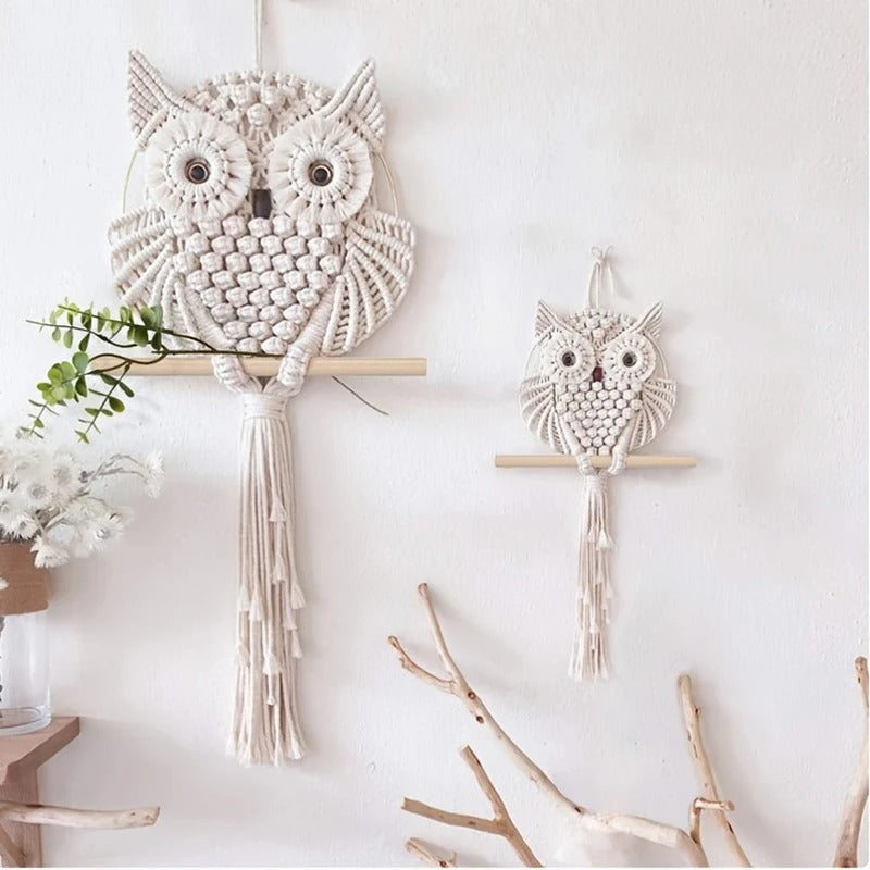 Deux tentures murales en macramé-hibou couleur crème, une grande et une petite, sont exposées sur un mur blanc. Suspendues à des tiges en bois, elles sont idéales pour la décoration intérieure et s'harmonisent parfaitement avec des touches d'art bohème comme des fleurs séchées.