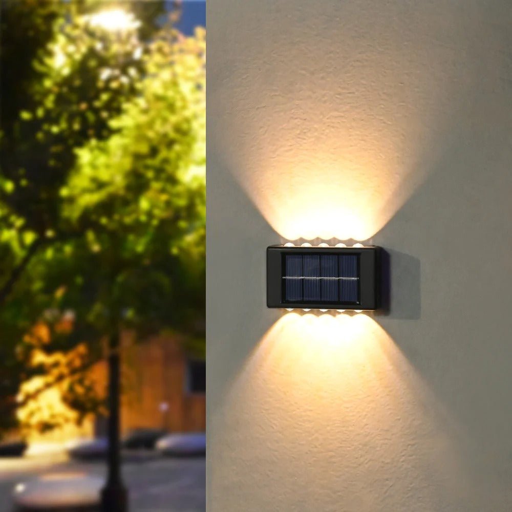 La Lampe solaire jardin haut de gamme /Del-Sol™ émet une lueur chaleureuse à double faisceau, éclairant le dessus et le dessous d'un mur extérieur ; parfait pour enrichir l’éclairage solaire extérieur avec une touche élégante, au milieu d’un décor d’arbres et de bâtiments.
