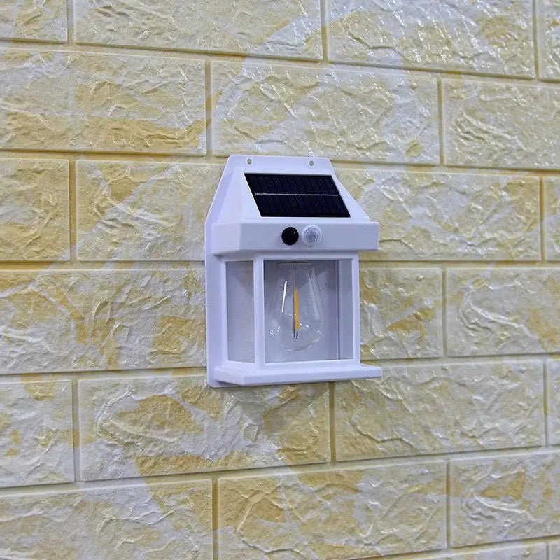 La Lampe solaire murale-Solar-LED™, une applique murale d'extérieur blanche avec capteur, est fixée sur un mur en briques jaunes texturées. Elle est dotée d'un petit panneau solaire sur le dessus et d'un couvercle transparent qui protège l'ampoule à l'intérieur.