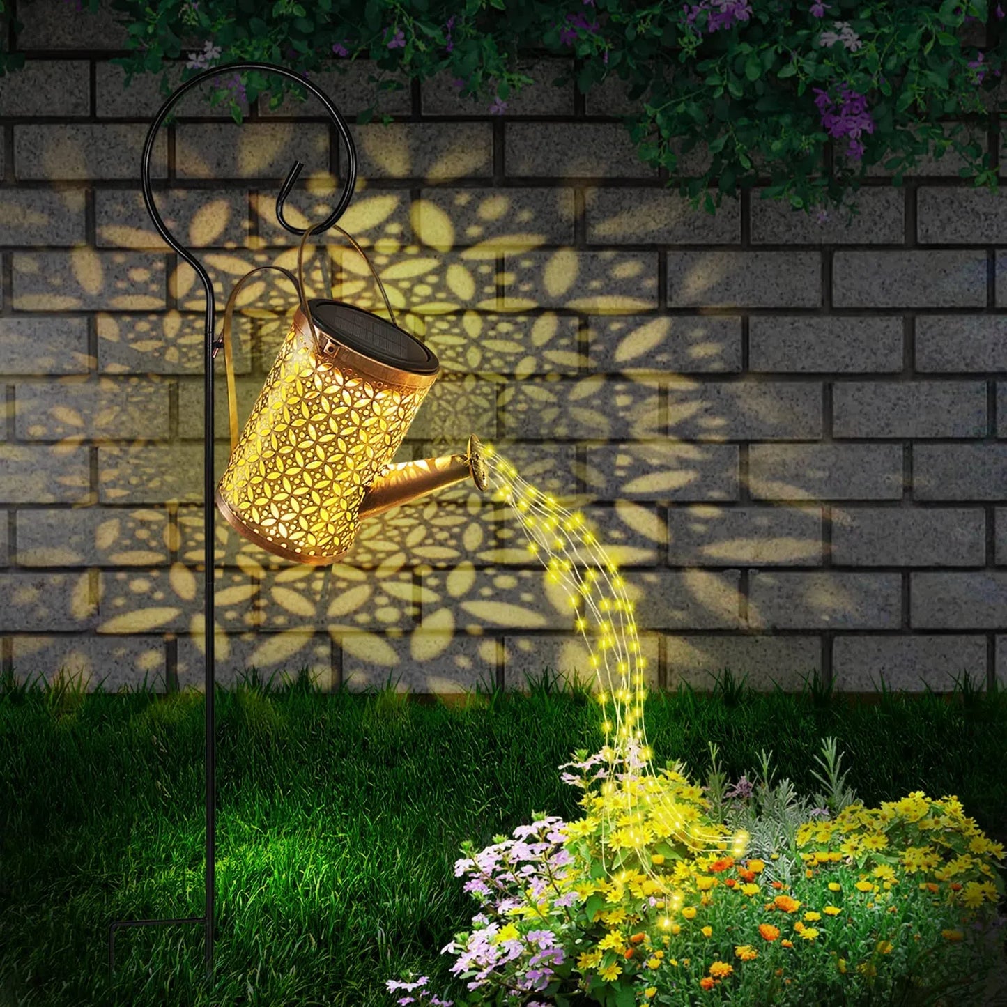 La Lampe solaire pour le jardin-Arrosoir, suspendue à un crochet, diffuse une lumiÚre jaune chaleureuse et projette des motifs floraux décoratifs sur le mur de briques tout en illuminant les fleurs en dessous la nuit.