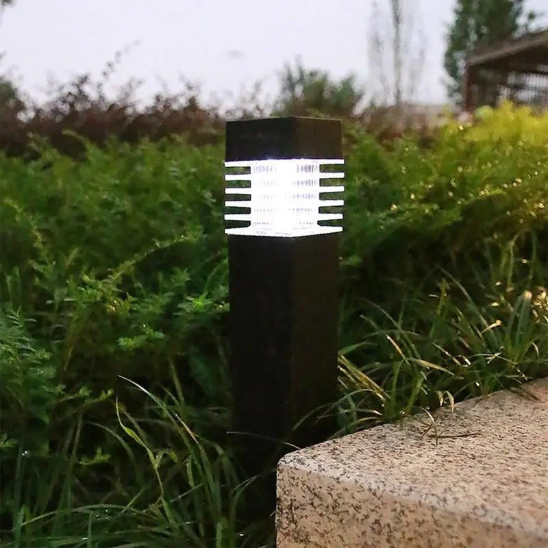 Une lampe de jardin extérieure noire émettant une douce lueur se dresse au milieu dune herbe verte luxuriante. Larrière-plan présente un feuillage flou et un chemin de pierre, suggérant un cadre de jardin tranquille.