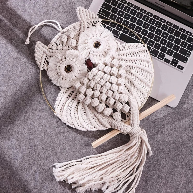 Le Macramé-Hibou, un hibou fait main avec des nœuds et des pompons complexes sur un perchoir en bois, repose sur une surface grise à côté d'un ordinateur portable - un accent idéal pour ajouter un charme bohème à votre décoration intérieure.