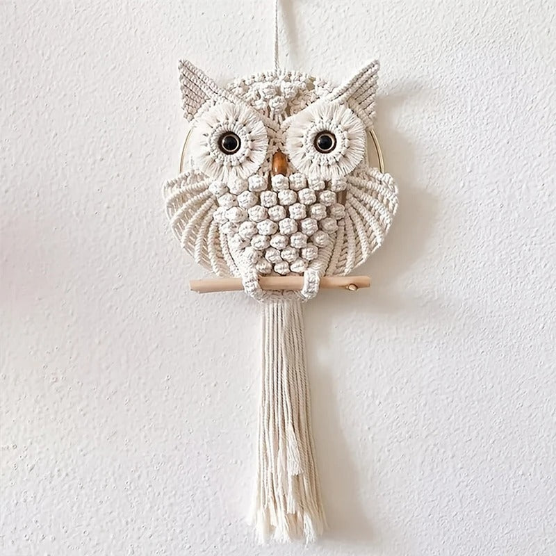 Le Macramé-Hibou, une tenture murale blanche faite à la main avec des nœuds et des pompons détaillés sur une cheville en bois, rehausse votre décoration intérieure avec une touche artistique bohème.