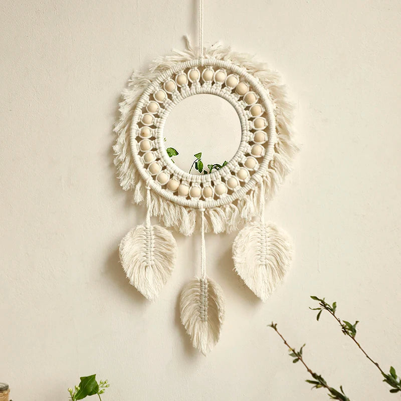 Un miroir rond est délicatement suspendu au centre d'une pièce murale en macramé blanc crème. Des cordons noués forment un motif rayonnant autour du miroir, agrémenté de perles de bois claires. Trois éléments décoratifs en forme de feuilles, également réalisés en macramé, pendent gracieusement sous le cercle central. L'ensemble évoque un style bohème et artisanal.