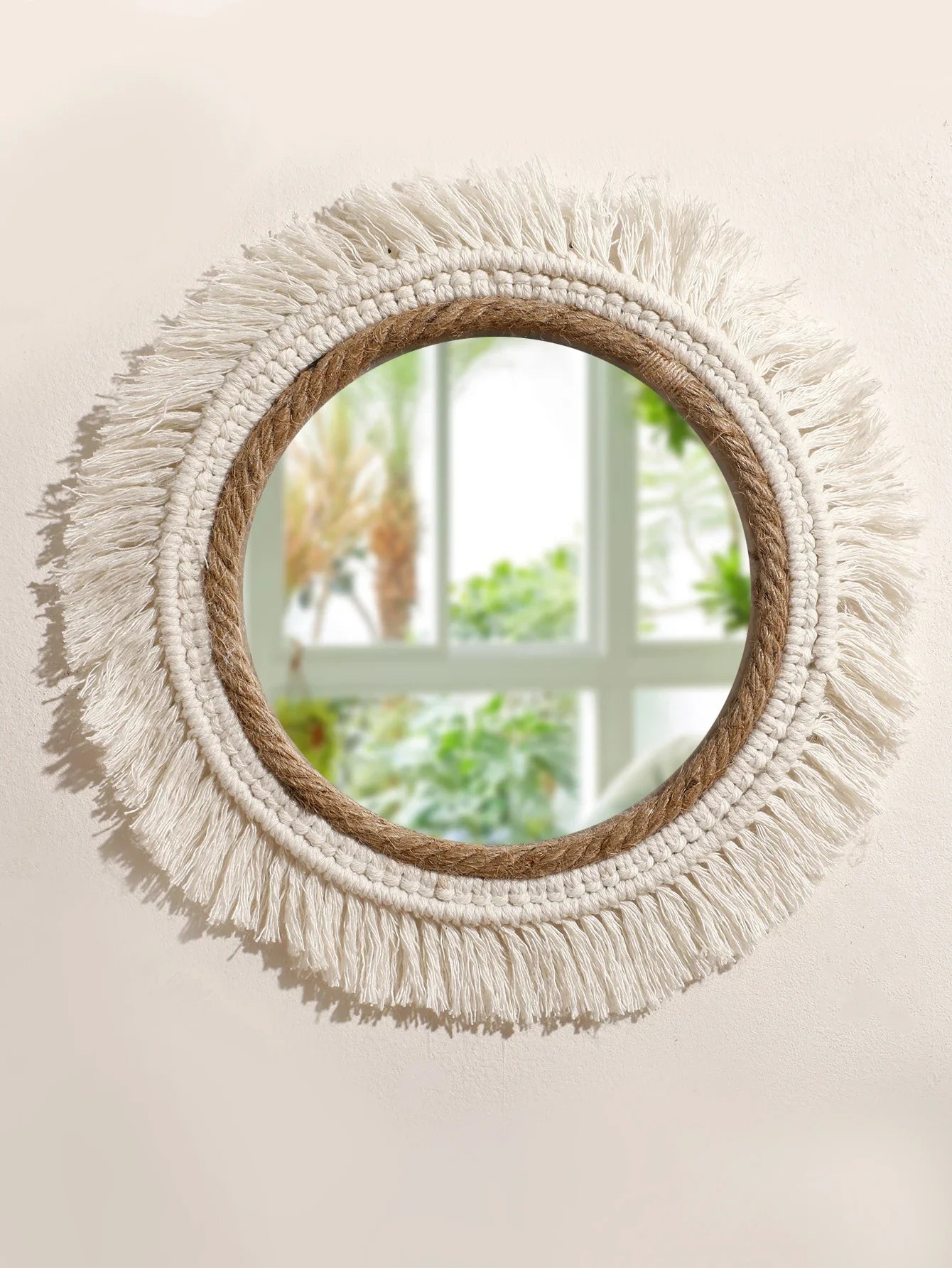 Un élégant miroir rond orné d'une bordure en corde naturelle est suspendu à un mur crème. Un délicat travail de macramé blanc cassé forme une auréole de franges autour du miroir, ajoutant une touche bohème et artisanale. Le reflet dans le miroir révèle une fenêtre lumineuse et des plantes verdoyantes, créant une atmosphère paisible et naturelle. L'ensemble capture la beauté simple et texturée du macramé.