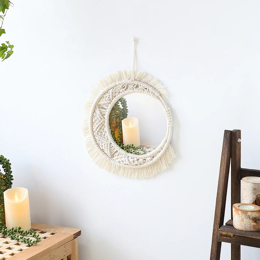 Un miroir rond suspendu au mur blanc est délicatement encadré par un ouvrage de macramé de couleur crème, formant un motif texturé et artistique. À l'intérieur du reflet du miroir, on aperçoit une bougie blanche allumée, ajoutant une douce lueur à la scène. Le travail de macramé, avec ses nœuds et ses franges, apporte une touche bohème et artisanale à cet objet décoratif.