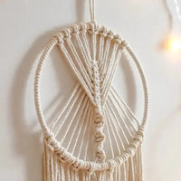 Un attrape-rêves circulaire en macramé de couleur crème est suspendu devant un mur blanc cassé.
Les cordons de macramé forment un motif géométrique complexe à l'intérieur du cercle.
De longues franges pendent du bas de l'anneau, créant une texture douce.
Une lumière douce et floue apparaît en arrière-plan à droite.