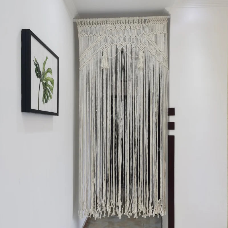 Un rideau macramé délicat, tissé en coton crème, orne une porte ou une fenêtre.
Le motif complexe présente des détails noués et des franges pendantes, ajoutant une texture bohème à la pièce.
La lumière filtre doucement à travers les fils, créant une ambiance chaleureuse et invitante.
Un tableau encadré d'une feuille verte est visible sur le mur blanc à gauche du rideau.