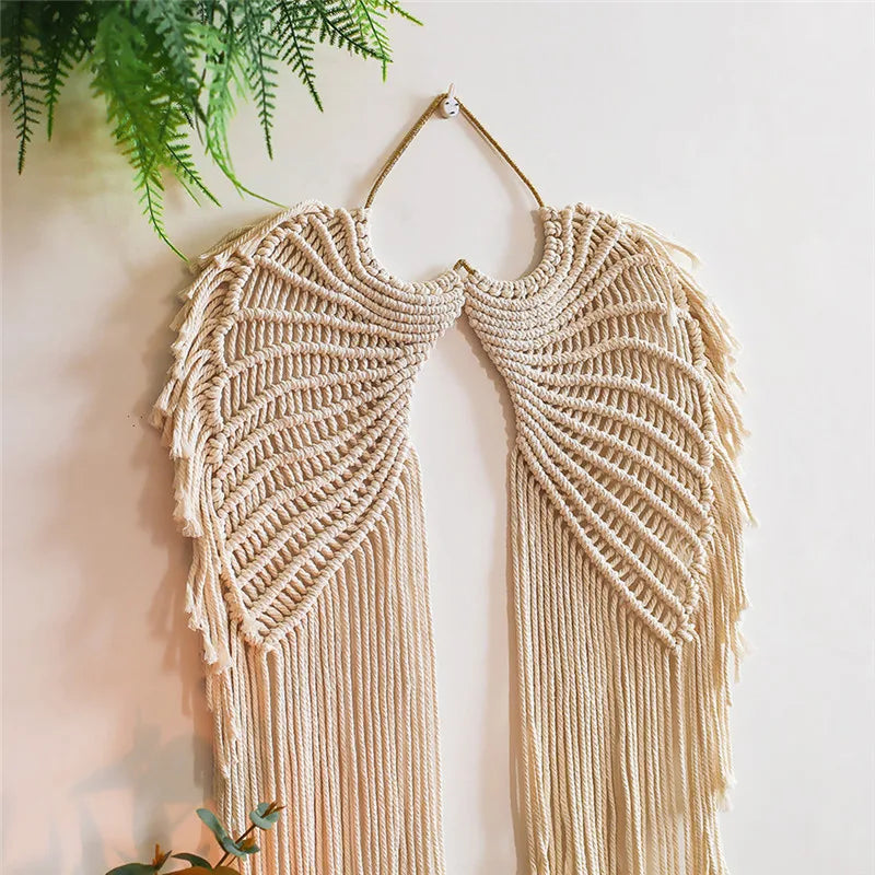 une décoration murale en macramé beige en forme d'ailes d'ange, suspendue sur un mur blanc. La texture complexe du macramé est bien visible, avec de longues franges pendantes. L'ensemble crée une ambiance bohÚme et artisanale, parfaite pour embellir un intérieur avec une touche de macramé.