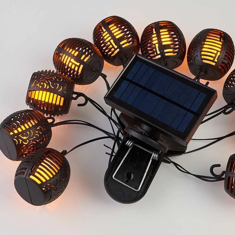 La guirlande extérieure solaire | Flamme est un ensemble de guirlandes lumineuses rondes noires de style lanterne avec des motifs découpés complexes et un panneau solaire central. Elle comprend un clip facile à attacher et émet une lueur orange chaude, offrant un effet flamme enchanteur pour votre espace.