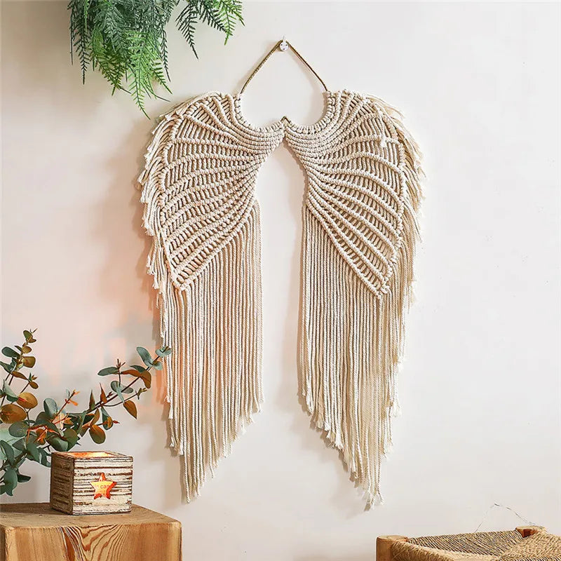 Cette photo présente une décoration murale en macramé beige en forme d'ailes d'ange, suspendue sur un mur blanc. La texture complexe du macramé est bien visible, avec de longues franges pendantes. L'ensemble crée une ambiance bohÚme et artisanale, parfaite pour embellir un intérieur avec une touche de macramé.