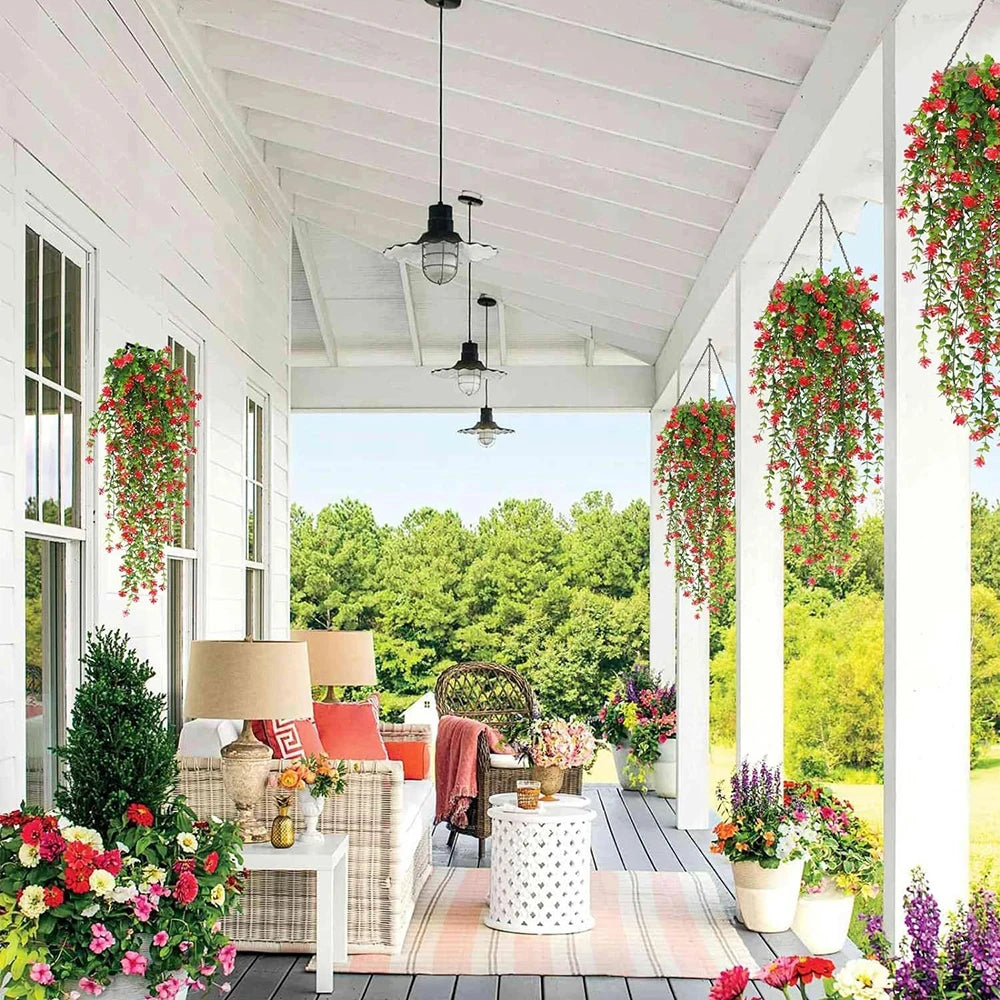 Un charmant porche aux murs blancs et aux colonnes présente des fleurs suspendues et en pot, des chaises en osier, une petite table, des lampes et les Plantes artificielles tombantes-Cascade Bohème pour une atmosphère relaxante donnant sur le jardin lumineux.