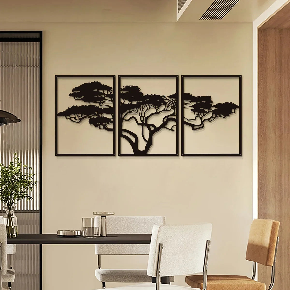 décoration murale en trois parties représentant un arbre de vie stylisé en métal noir sur un mur beige clair. La salle à manger moderne présente cette œuvre d'art unique au-dessus d'une table noire et de chaises aux tons neutres.