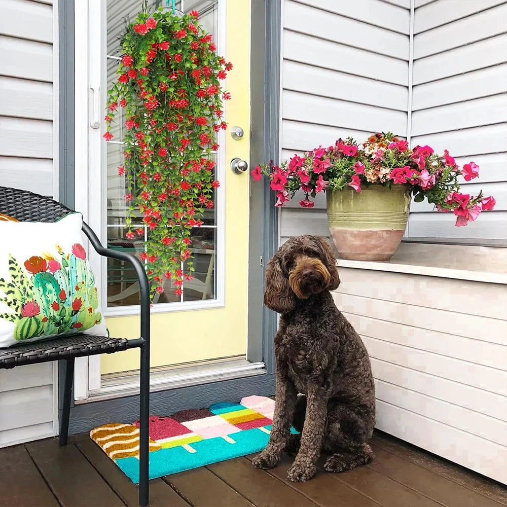 Un chien brun aux cheveux bouclés est assis sur un paillasson coloré près d'une porte jaune. Le revêtement gris du porche, la chaise avec son coussin et les plantes artificielles tombantes Cascade Bohème aux fleurs rose vif créent un décor végétal luxuriant et apaisant.