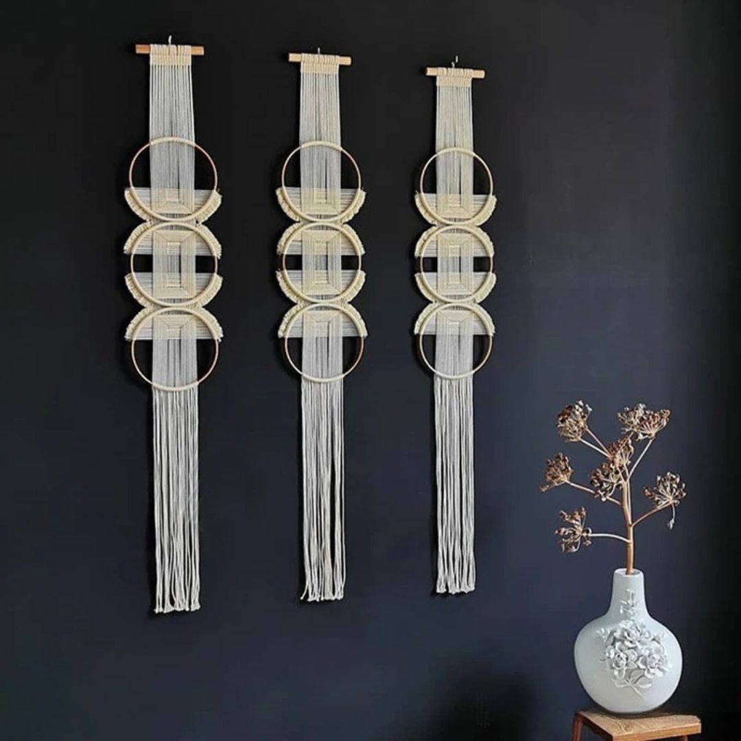Trois décorations murales en macramé de couleur crème ornent un mur bleu foncé. Chaque pièce est suspendue à une baguette en bois clair et présente un motif de cercles en métal entrelacés avec des cordes, terminées par de longues franges pendantes. Un vase blanc texturé contenant une branche séchée est posé sur un petit tabouret en bois sur le côté droit. L'ensemble crée une ambiance bohème et artisanale. Marcrame.