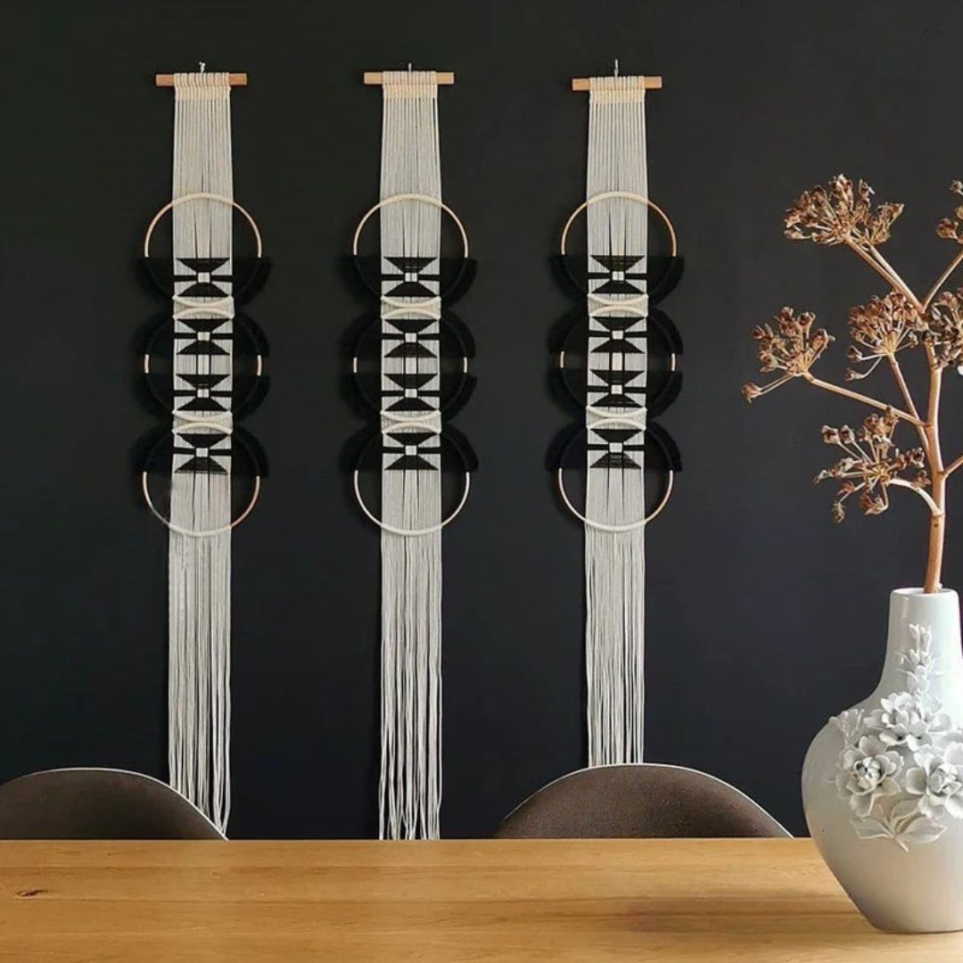 Trois décorations murales en macramé ivoire et noir, suspendues à des tiges en bois clair, ornent un mur sombre. Chaque pièce présente des motifs géométriques complexes et des cercles dorés, créant un effet visuel captivant. Sur une table en bois, un vase blanc texturé contient des fleurs séchées délicates. L'ensemble évoque une ambiance bohème chic et artisanale. #macrame