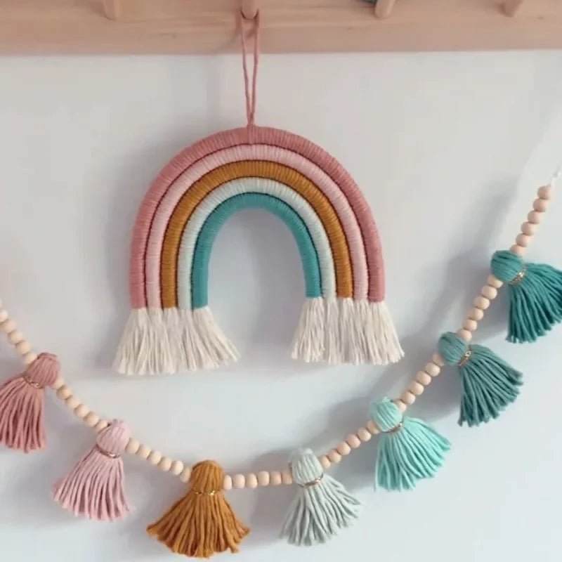 Un arc-en-ciel décoratif en macramé aux couleurs pastel, suspendu à un mur blanc. En dessous, une guirlande de perles de bois et de glands en fil de macramé dans des tons assortis de rose, ocre et turquoise. L'ensemble crée une ambiance douce et artisanale. La texture nouée du macramé est bien visible.