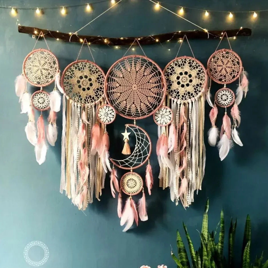 Un arrangement en macramé décoratif suspendu orne un mur bleu-vert, composé de plusieurs attrape-rêves de différentes tailles. Chaque cercle est délicatement tissé avec des motifs de macramé en tons de pêche et crème, agrémenté de plumes douces et de rubans flottants. Une branche naturelle sert de support supérieur, illuminée par une guirlande lumineuse chaleureuse.
