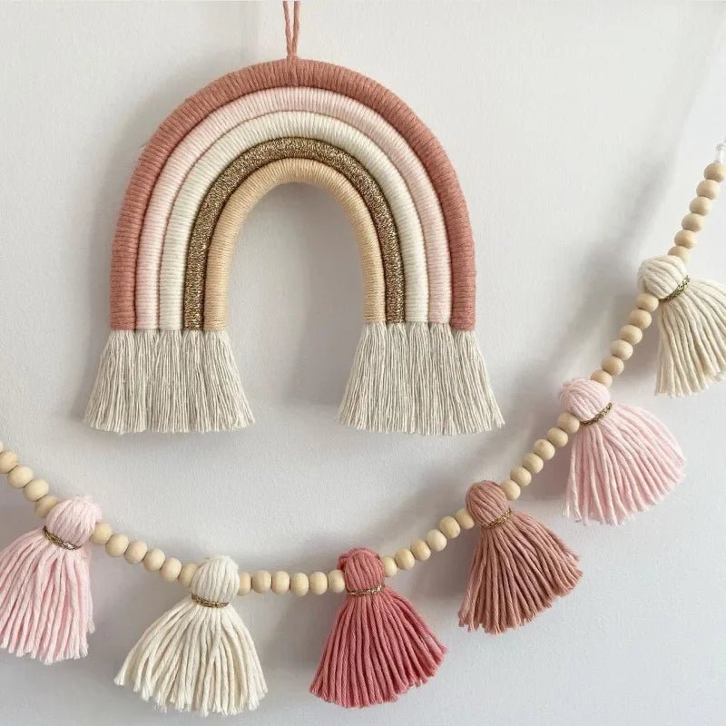 Un bel arc-en-ciel décoratif en macramé orne le mur. Ses bandes de couleurs douces, allant du rose poudré au beige scintillant, sont suspendues par une cordelette. En dessous, une guirlande de perles de bois alterne avec des pompons en macramé dans des tons similaires, créant une ambiance chaleureuse et artisanale.