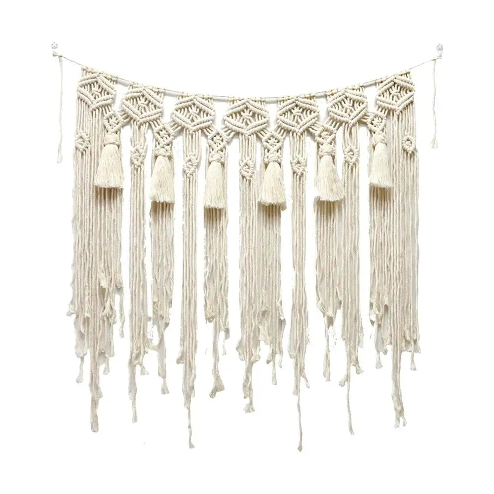 Un délicat rideau macramé couleur crème, suspendu par une fine baguette de bois. Des motifs géométriques complexes ornent la partie supérieure, tandis que d'épaisses franges torsadées et des glands pendants ajoutent une texture bohème. La lumière douce révèle les détails noués de ce Rideau macramé artisanal.