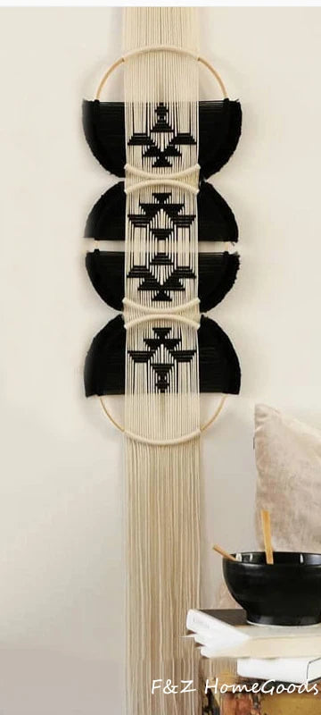 Un élégant macramé mural beige et noir orne un mur clair. Le motif central, tissé avec soin, est encadré par des cercles de bois clair. De longues franges beiges tombent gracieusement du bas de cette décoration marcrame, ajoutant une texture douce à l'ensemble.