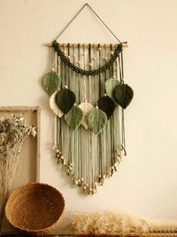 Un macramé décoratif suspendu à un mur beige clair présente un motif de feuilles vertes et beiges. Des perles de bois clair parsèment les cordes et ornent le bas de la pièce. L'art mural crée une ambiance bohème et naturelle. Marcame.