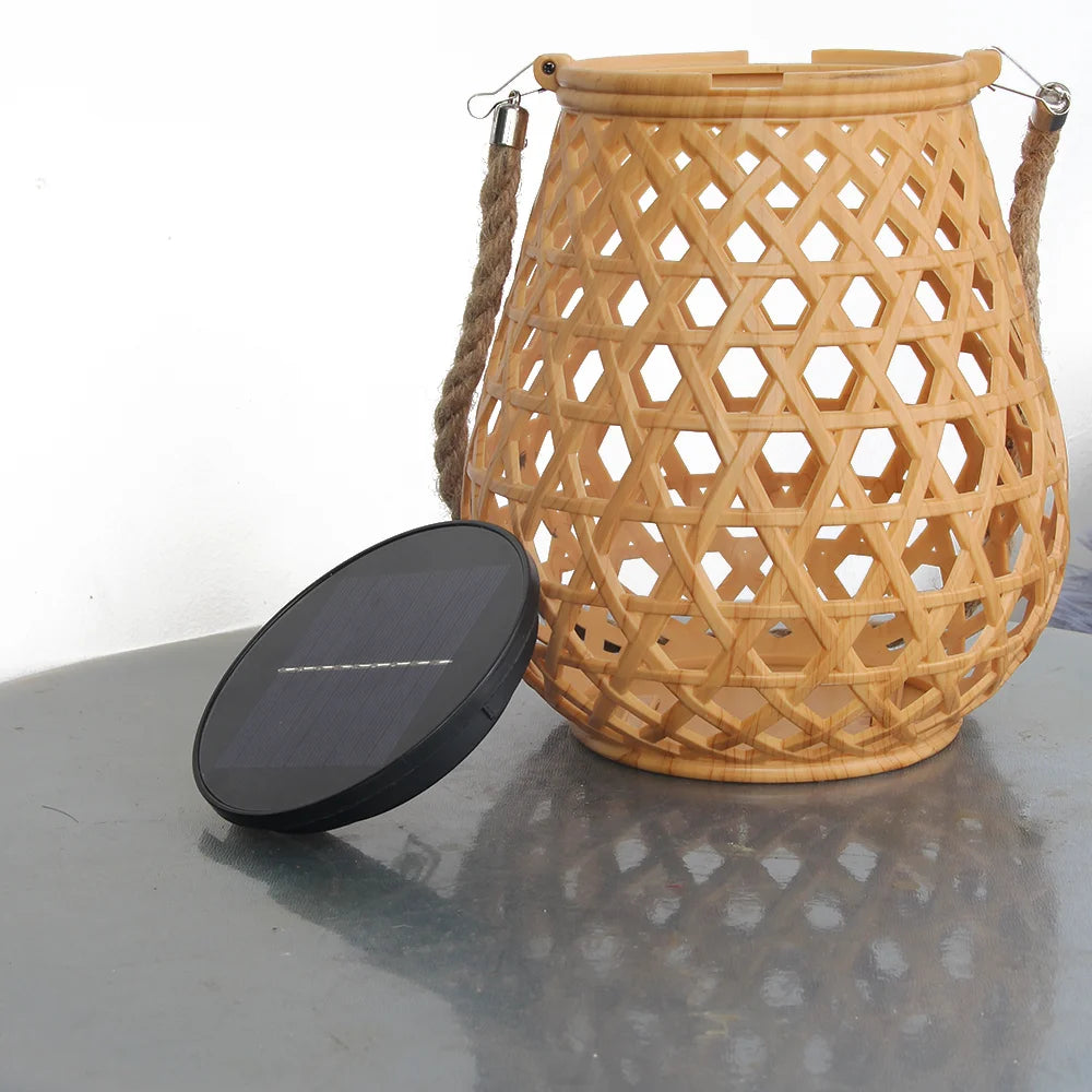 Une Lampe solaire de jardin haut de gamme en Bambou Tissé avec une poignée en corde repose sur une surface réfléchissante ; son panneau solaire noir et circulaire repose à proximité contre un mur blanc uni, soulignant sa conception pour l'éclairage écologique.