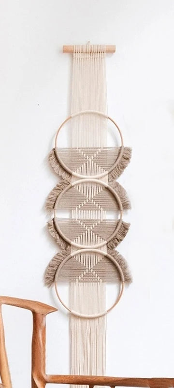 Une décoration murale en macramé beige et ficelle écrue est suspendue à un bâton en bois clair. Trois cercles ornés de motifs géométriques complexes en macramé beige se superposent le long d'une bande verticale de ficelle écrue. Des franges beiges pendent des parties inférieures des cercles, ajoutant une texture douce à cette pièce de marcrame décorative. L'ensemble est accroché sur un mur blanc uni.