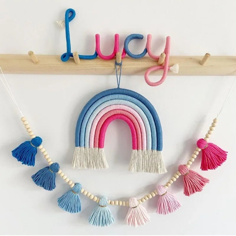 Une décoration murale enfantine présente un arc-en-ciel en macramé aux couleurs vives suspendu sous le prénom "Lucy" écrit en fil coloré. Une guirlande de glands bleus et roses, séparés par des perles en bois, complète cet arrangement décoratif suspendu à un support en bois sur un mur blanc. Cet ensemble crée une ambiance douce et artisanale.
