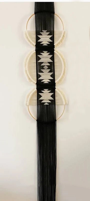 Une pièce de macramé décorative est suspendue au mur blanc. Elle est composée de trois cercles en bois, chacun partiellement rempli de cordes de couleur crème formant des demi-lunes. Une section centrale de macramé noir descend verticalement, ornée de motifs géométriques blancs. De longues franges noires pendent du bas de cette section, créant une texture visuelle intéressante. #macrame