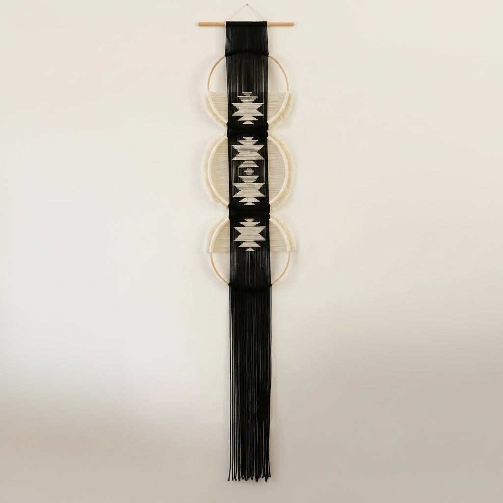 Une pièce de macramé décorative est suspendue au mur blanc. Elle est composée de trois cercles en bois, chacun partiellement rempli de cordes de couleur crème formant des demi-lunes. Une section centrale de macramé noir descend verticalement, ornée de motifs géométriques blancs. De longues franges noires pendent du bas de cette section, créant une texture visuelle intéressante. #macrame