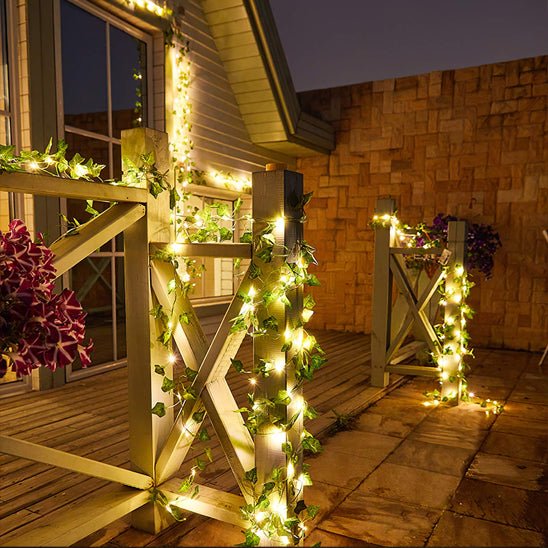 La terrasse extérieure est dotée de balustrades en bois ornées de lierre et d'une guirlande solaire extérieure Lueur de bonheur, qui brille doucement. Deux jardinières aux fleurs violettes sont posées sur le sol carrelé, tandis qu'un mur de pierre et une partie d'une maison en arrière-plan créent une ambiance de soirée chaleureuse, flexible et économique.
