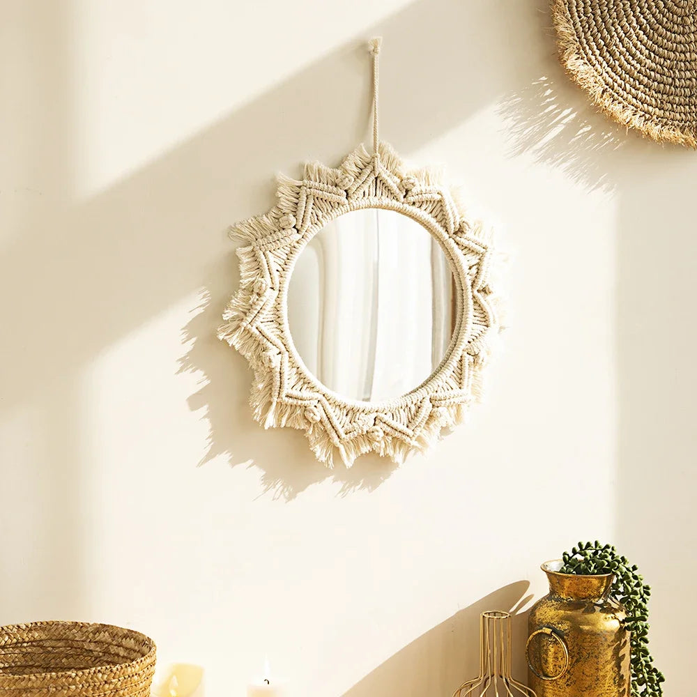 Un miroir rond est délicatement encadré par un travail de macramé complexe en forme d'étoile. Les franges de coton crémeux rayonnent autour du verre réfléchissant, capturant la lumière douce de la pièce. Suspendu à un mur blanc, il projette de subtiles ombres, ajoutant une touche chaleureuse et artisanale à la décoration.
