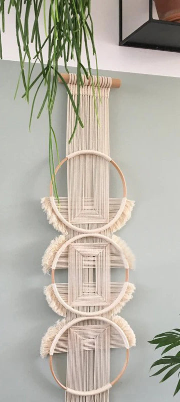 Décoration murale en macramé beige suspendue à un mur bleu clair. La pièce de macramé est composée de trois cercles en bois concentriques, ornés de motifs géométriques en corde et de franges texturées. La mention "marcrame" est visible dans le design de l'œuvre.