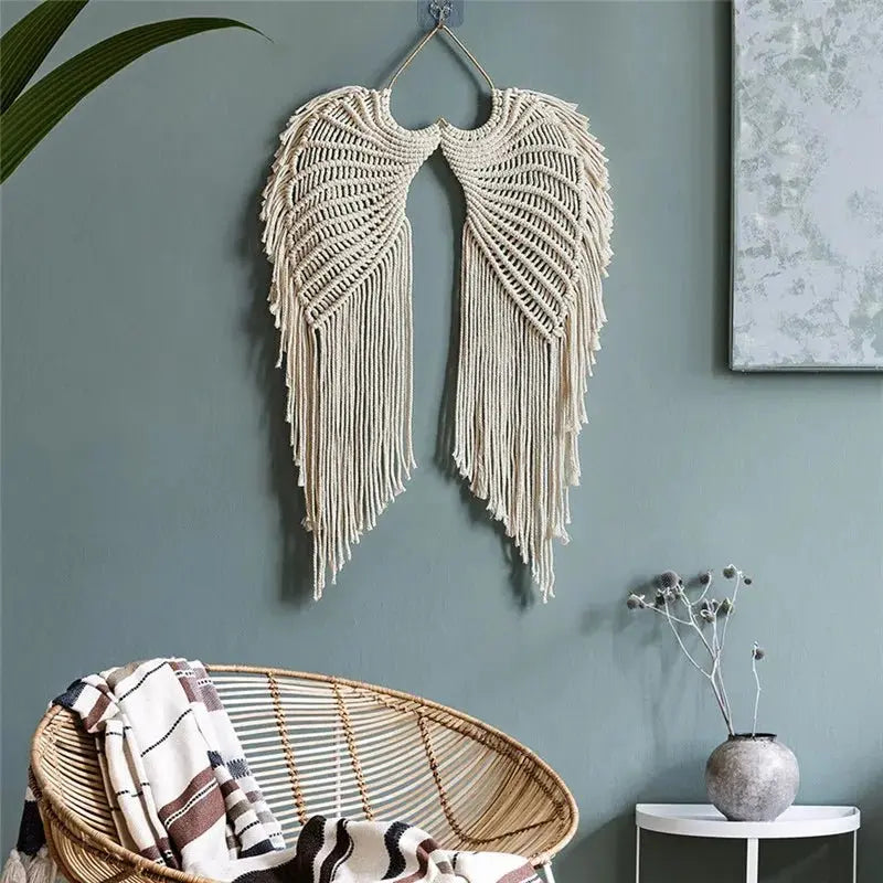 Une décoration murale en macramé représentant des ailes d'ange est suspendue sur un mur bleu-vert. La pièce artisanale en corde de coton apporte une touche bohème à l'intérieur. Un fauteuil en rotin et une petite table complètent le décor.
