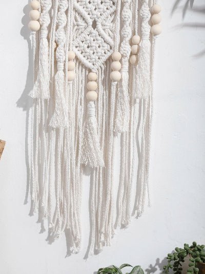 délicat ouvrage de macramé en coton écru orne un mur blanc texturé. Le motif central présente un losange complexe, encadré de cordes nouées et de perles de bois clair. De longues franges pendantes ajoutent une touche bohème à cette pièce artisanale. Des ombres douces et des fragments de plantes vertes en bas complètent le tableau.