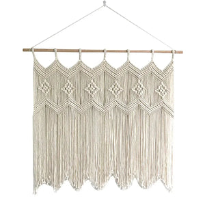 Un rideau en macramé de couleur crème, suspendu par une corde à une tringle en bois clair, est centré sur un fond blanc. Le motif complexe du macramé comprend des losanges centraux et des détails noués sur toute la surface. Le bas du rideau présente une bordure festonnée de franges tombantes.