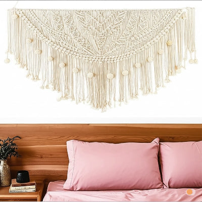 Un gros plan présente une magnifique tenture murale en macramé blanc cassé ornant le mur au-dessus d’un lit avec une literie et des oreillers roses. Le tissage complexe forme un motif semi-circulaire avec des franges et des sphères décoratives le long du bord inférieur. En dessous, on aperçoit la tête de lit en bois ainsi qu’une table de nuit portant des livres et un vase de verdure.