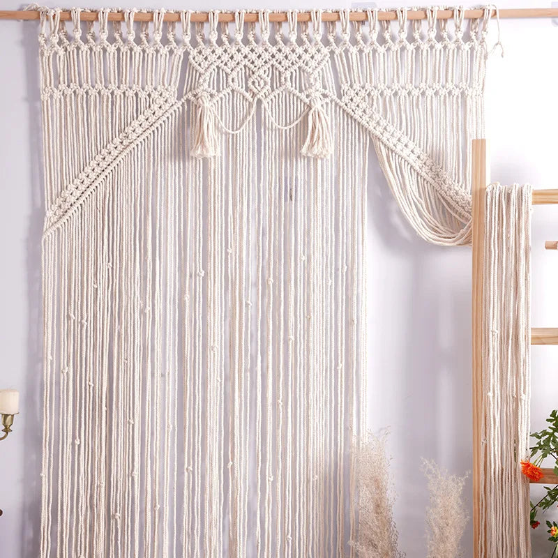 Un rideau macramé ivoire complexe, suspendu à une tringle en bois naturelle.
Le motif présente un panneau supérieur décoratif avec des détails noués et des franges pendantes.
De longs brins de macramé forment le corps du rideau, créant une texture aérée et bohème.
Une section du rideau est tirée sur le côté, révélant un fond blanc uni.
