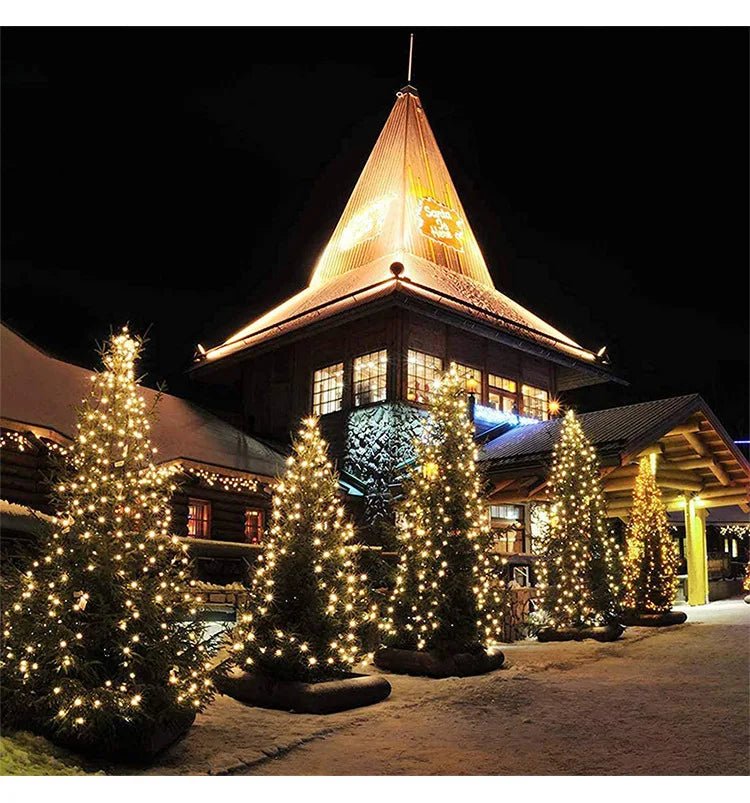 Une scène nocturne festive met en scène des sapins de Noël illuminés de lumières blanches chaudes et de LED ultra-vives. Un bâtiment en bois au toit pyramidal brille doucement, tandis que la neige recouvre le sol, ajoutant au charme hivernal. Vivez cette ambiance avec Guirlande solaire extérieur | SolarSpark.