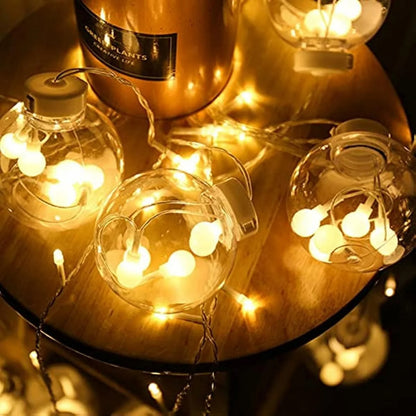 Des guirlandes lumineuses blanc chaud et des ampoules rondes en verre sont disposées sur une surface en bois. Un pot de fleurs de couleur cuivre marqué « PLANTS » est partiellement visible. La Guirlande Solaire Extérieur Puissante | Lueur Puissante crée une lueur chaleureuse et ambiante pour un cadre extérieur enchanteur.
