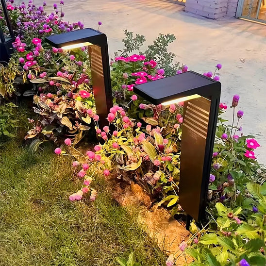 Dans un jardin luxuriant aux fleurs roses et violettes, deux lampes noires épurées Lampe solaire extérieur | Écolo projettent une lumière chaleureuse sur les fleurs et le feuillage. Des pavés et un muret ajoutent à lambiance écologique.