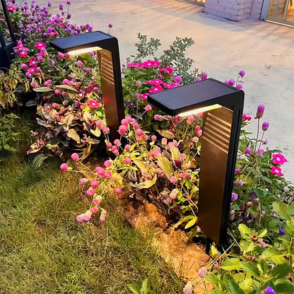 Dans un jardin luxuriant aux fleurs roses et violettes, deux lampes noires épurées Lampe solaire extérieur | Écolo projettent une lumière chaleureuse sur les fleurs et le feuillage. Des pavés et un muret ajoutent à lambiance écologique.